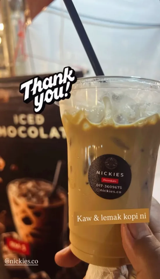 Customer holding a Nickies Premium iced coffee in front of the Iced Chocolate banner, with the note “Kaw & lemak kopi ni”