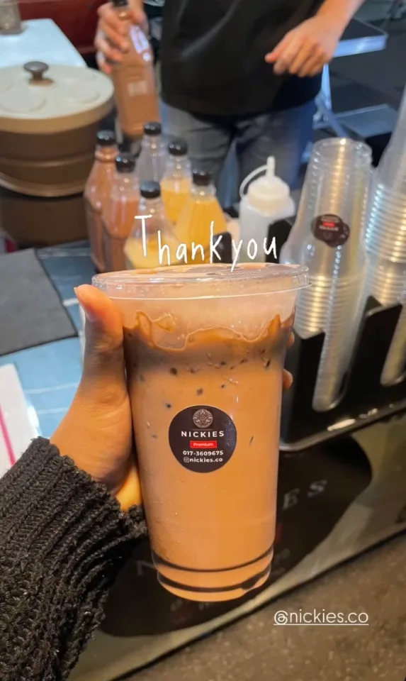 Customer holding a Nickies iced chocolate with caramel drizzle at the stall counter, with bottles and stacked cups behind